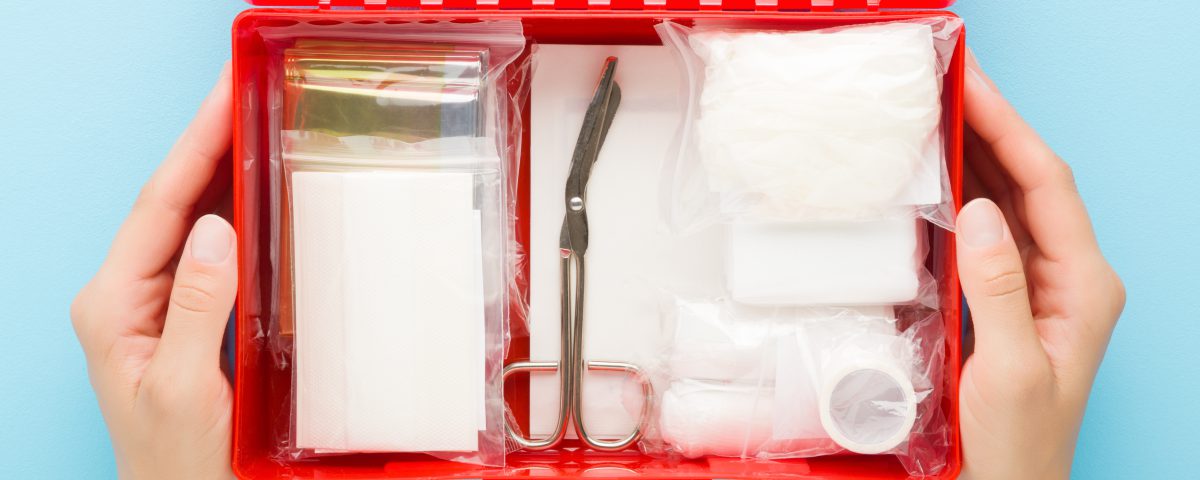Young adult woman hands holding opened red first aid kit box with different medical accessories in packages on light blue table background. Pastel color. Closeup. Point of view shot. Top down view.