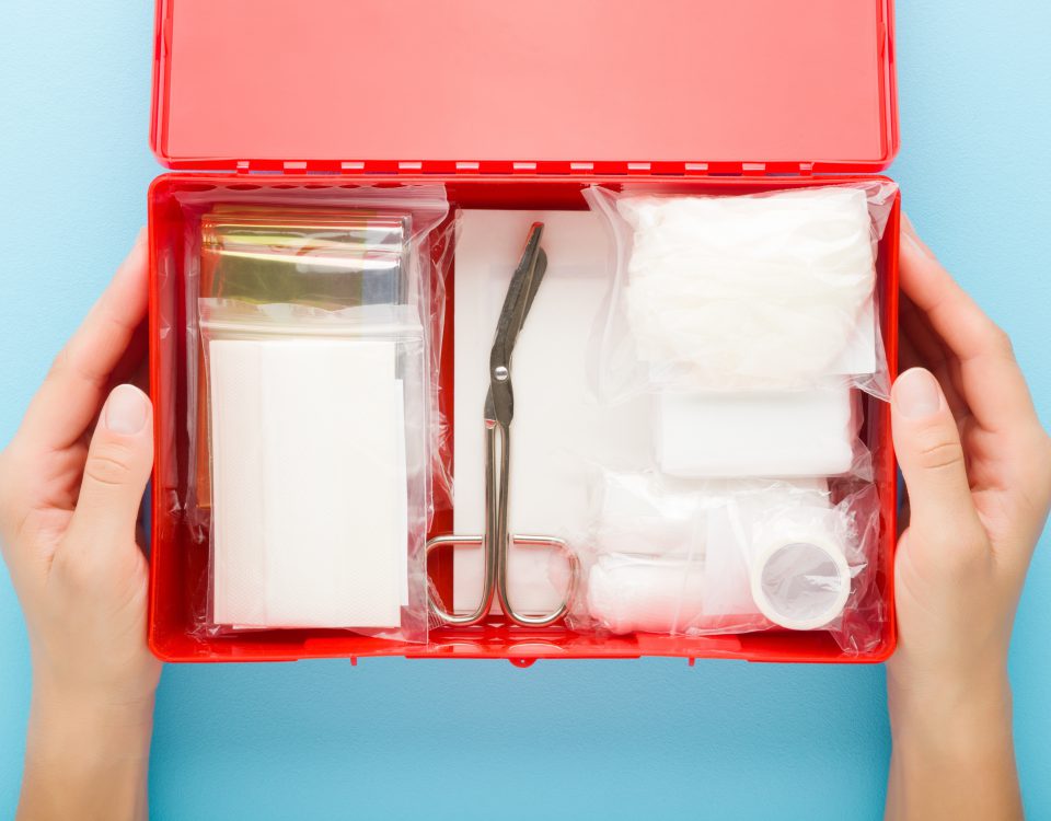 Young adult woman hands holding opened red first aid kit box with different medical accessories in packages on light blue table background. Pastel color. Closeup. Point of view shot. Top down view.